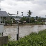 Stasiun sensor pemantau banjir bertenaga surya di tepi sungai dengan latar permukiman dan langit mendung